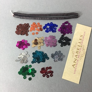 May include: Assortment of 12 colors of round sequins, each pile containing approximately 50 sequins.  The sequins are arranged in rows of three.  A brown, flat, beaded cord is included.  The image also includes a business card for Anabelles Vintage Beads & Findings.