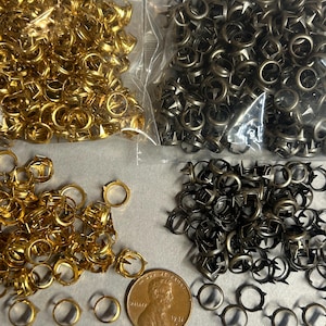 May include: An assortment of metal rivets and fasteners in gold and gunmetal colors. The image shows various shapes and sizes of rivets, including some with ring-like designs. A US penny is included for size reference.