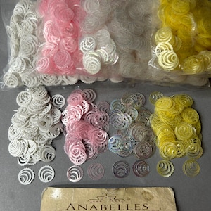 May include: Four bags of iridescent plastic rose-shaped beads in pink, white, iridescent, and yellow. The bags are open and the beads are spilling out. There is a sign that says "Anabelles Vintage Beads & Findings" with contact information and hours of operation.