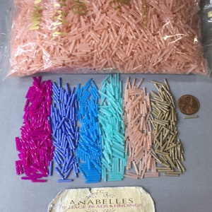 Op de afbeelding: Een doorzichtige plastic zak gevuld met roze zaadkralen. Daaronder bevinden zich geordende rijen zaadkralen in tinten fuchsia, blauw, turquoise, perzik en bruin. Een munt ligt naast de bruine kralen. Onderaan een visitekaartje.