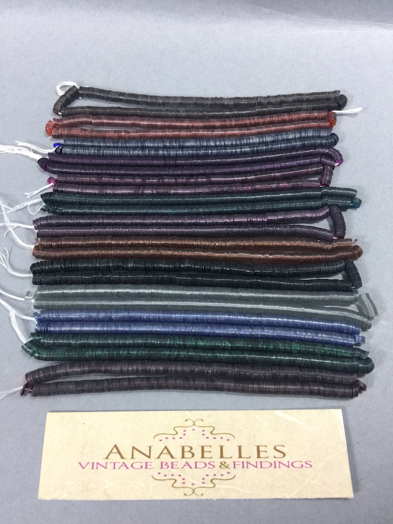 May include: A collection of 18 strands of iridescent, flat, round, plastic beads in various colors, including black, brown, purple, green, and gray. The beads are arranged in rows and are all the same size. The beads are labeled "Anabelles Vintage Beads & Findings".