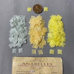 May include: Three piles of flower-shaped sequins in white, cream, and yellow. The sequins are arranged in a circle around a penny. A business card for Anabelles Vintage Beads & Findings is below the sequins.