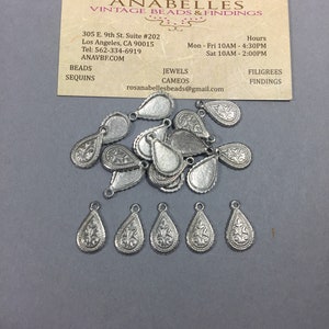May include: A pile of silver teardrop-shaped charms with intricate floral designs. The charms have a loop at the top for attaching to jewelry or other crafts.
