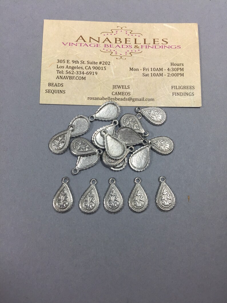 May include: A pile of silver teardrop-shaped charms with intricate floral designs. The charms have a loop at the top for attaching to jewelry or other crafts.