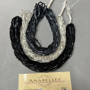 May include: Two strands of black and silver tube beads, each with a white thread attached. The beads are arranged in a circle on a white surface. A business card for Anabelles Vintage Beads & Findings is in the lower right corner of the image. The card includes the business address, phone number, website, and hours of operation.