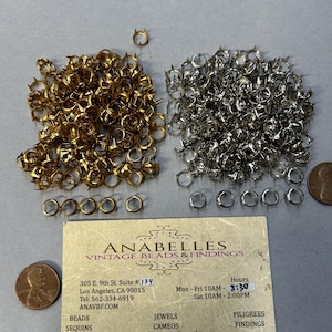 May include: A close-up image of two piles of gold and silver metal crown settings. The settings are small and have a pointed crown design. The image also includes a business card for Anabelles Vintage Beads & Findings, with their address, phone number, and website. The business card also lists their hours of operation: Monday to Friday 10am to 3:30pm and Saturday 10am to 2:00pm.
