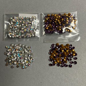 Könnte beinhalten: Vier kleine Plastiktüten gefüllt mit Strasssteinen. Zwei Tüten enthalten schillernde Strasssteine, die anderen beiden eine Mischung aus goldenen und lila Strasssteinen. Die Strasssteine sind auf einer grauen Oberfläche verstreut.