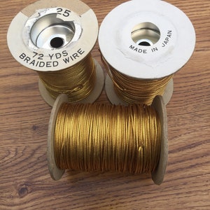 May include: Three spools of gold braided wire on a wooden surface. One spool has the text "72 YDS BRAIDED WIRE" and the number "25". Another spool has the text "MADE IN JAPAN". The wire is tightly wound around cardboard spools.