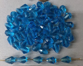 May include: A pile of turquoise blue glass beads with a faceted, teardrop shape. The beads are approximately the size of a US penny. A string of the beads is shown in the foreground.