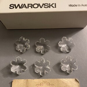 May include: Six clear crystal snowflake charms with a Swarovski Crystal box. The box is white with black text that reads "CRYSTAL SWAROVSKI Made in Austria".
