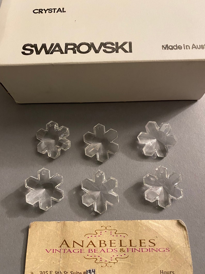 May include: Six clear crystal snowflake charms with a Swarovski Crystal box. The box is white with black text that reads "CRYSTAL SWAROVSKI Made in Austria".
