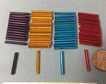 May include: Assortment of four colors of glass beads, each color in a stack of approximately 20 beads. The beads are approximately 1 inch long and have a rectangular shape. The colors are purple, orange, blue, and red. A single bead of each color is shown below the stacks.