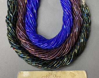 May include: Three strands of glass beads, one blue, one purple, and one green, with iridescent finishes. The beads are all the same size and shape, and they are arranged in a loose circle. The beads are for sale at Anabelles Vintage Beads & Findings, 305 E. 9th St. Suite #134, Los Angeles, CA 90015. Tel: 562-334-6919. ANAVBA.COM. Hours: Mon - Fri 10AM-3:30PM, Sat 10AM - 2:00PM.