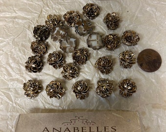 May include: A collection of gold-toned metal rose-shaped embellishments. The embellishments are arranged on a white surface. The image also includes a business card for Anabelles Vintage Beads & Findings.