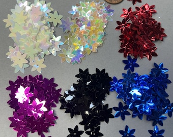 Puede incluir: Una colección de pequeñas lentejuelas con forma de flor de seis pétalos en blanco iridiscente, rojo metálico, morado, negro y azul. Se incluye una moneda de cobre para mostrar la escala y el tamaño relativo de las lentejuelas.