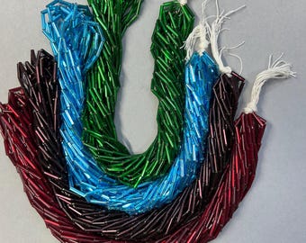 May include: Four strands of long, cylindrical glass bugle beads in vibrant colors including emerald green, bright blue, deep purple, and dark red. Each strand is secured with a white string tied at the top, showcasing a glossy, reflective finish.