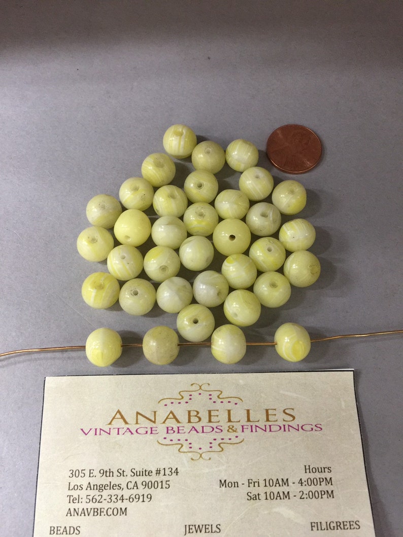 Op de afbeelding: Een stapel van 25 gele en witte glazen kralen met een witte draad erdoorheen. De kralen zijn rond en hebben een swirl-patroon. Een visitekaartje voor Anabelles Vintage Beads & Findings bevindt zich in de rechterbenedenhoek van de afbeelding.