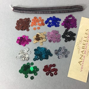 May include: Assortment of 12 colors of round sequins, each color in a separate pile. The sequins are metallic and have a shiny finish. A brown, flat, rectangular piece of material is also included in the image.