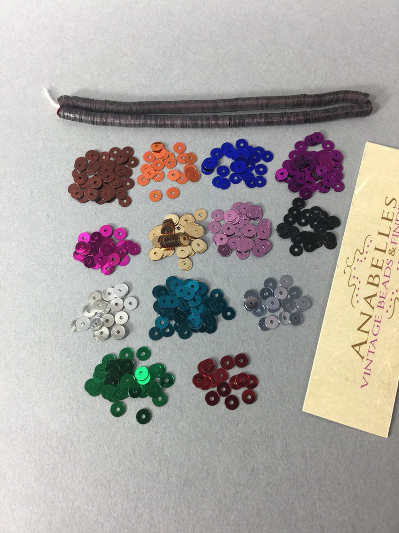 May include: Assortment of 12 colors of round sequins, each color in a separate pile. The sequins are metallic and have a shiny finish. A brown, flat, rectangular piece of material is also included in the image.