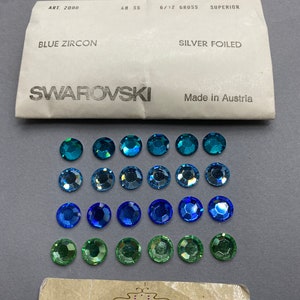 May include: A close-up of 24 assorted Swarovski crystal rhinestones in shades of blue and green. The rhinestones are arranged in three rows of eight. The rhinestones are packaged in a white envelope with the text "ART. 2000", "48 SS", "6/12 GROSS", "SUPERIOR", "BLUE ZIRCON", "SILVER FOILED", "SWAROVSKI", and "Made in Austria".