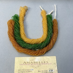 May include: Three spools of seed beads, one gold, one green, and one light brown. The spools are arranged in a U shape. The image includes the business name "ANABELLES VINTAGE BEADS & FINDINGS" and contact information.