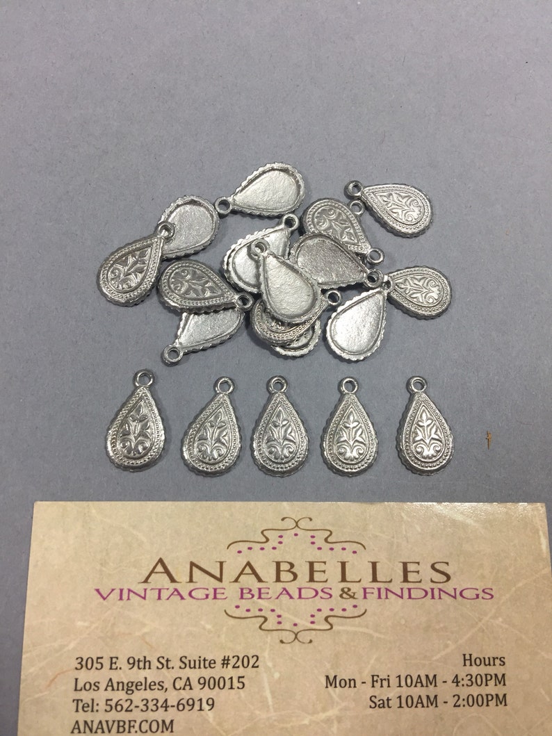 May include: A group of 15 silver teardrop-shaped metal charms with a decorative floral design. The charms have a loop at the top for attaching to jewelry or other crafts.