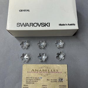 May include: A box of clear crystal snowflake beads from Swarovski. The box is white with black text that reads "ART. 6707", "25 MM", "2/12 GROSS", "SPECIAL", "CRYSTAL", "SWAROVSKI", and "Made in Austria". There are six clear crystal snowflake beads scattered around the box.