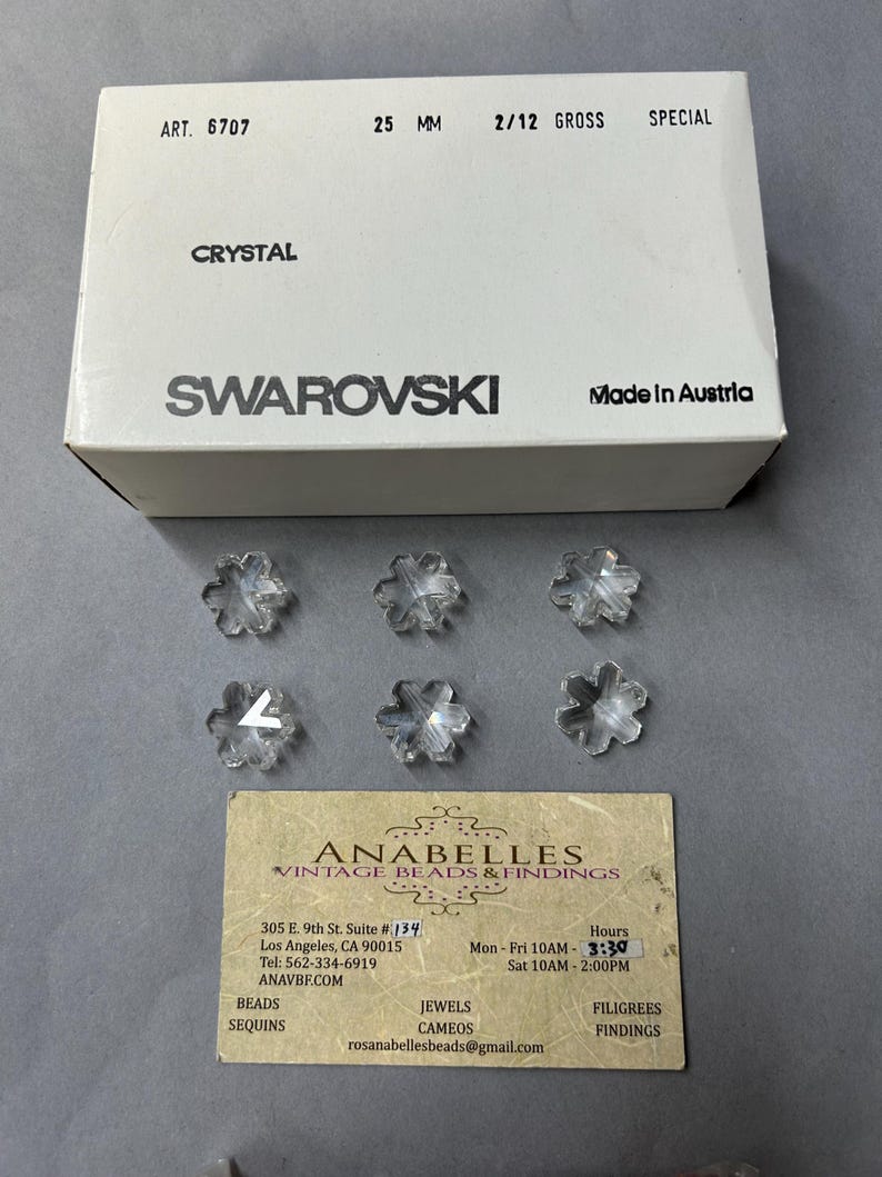 May include: A box of clear crystal snowflake beads from Swarovski. The box is white with black text that reads "ART. 6707", "25 MM", "2/12 GROSS", "SPECIAL", "CRYSTAL", "SWAROVSKI", and "Made in Austria". There are six clear crystal snowflake beads scattered around the box.