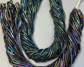 May include: Two strands of iridescent bugle beads featuring a metallic finish with shifting hues of deep blue, purple, and green. The long, cylindrical glass beads are strung on white thread, creating a reflective, multi-colored appearance.