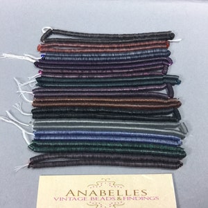 May include: A stack of 15 strands of iridescent glass beads in various colors, including black, brown, purple, green, and blue. The beads are arranged in a neat stack, with the ends of each strand visible. The beads are all the same size and shape, and they have a slightly textured surface. The beads are arranged on a white surface, and there is a small card with the text "ANABELLES VINTAGE BEADS & FINDINGS" in the bottom right corner of the image.