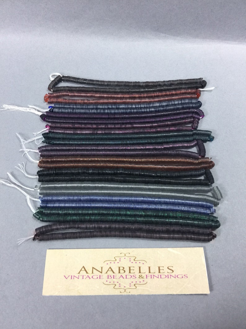 May include: A stack of 15 strands of iridescent glass beads in various colors, including black, brown, purple, green, and blue. The beads are arranged in a neat stack, with the ends of each strand visible. The beads are all the same size and shape, and they have a slightly textured surface. The beads are arranged on a white surface, and there is a small card with the text "ANABELLES VINTAGE BEADS & FINDINGS" in the bottom right corner of the image.