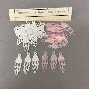 May include: A collection of white and pink plastic leaf-shaped embellishments. The embellishments have a cut-out design and are packaged with a label that reads "Registered Trade Mark - Made in France".