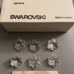 May include: Six clear crystal snowflake charms, each measuring 25 mm, packaged in a white box with the text "ART. 6707", "25 MM", "2/12 GROSS", "SPECIAL", "CRYSTAL", "SWAROVSKI", and "Made in Austria".