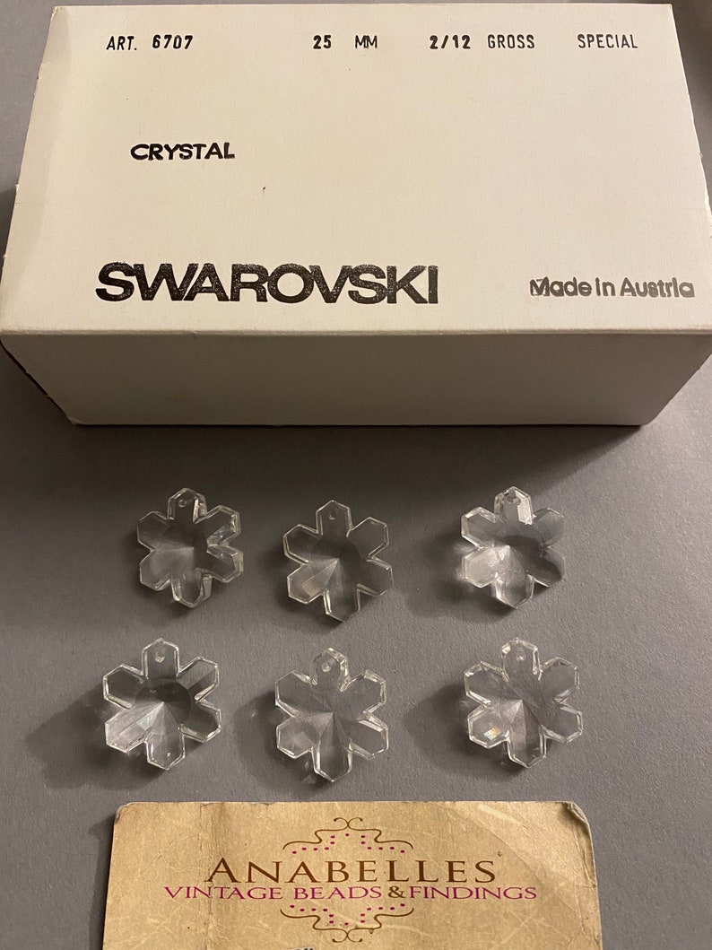 May include: Six clear crystal snowflake charms, each measuring 25 mm, packaged in a white box with the text "ART. 6707", "25 MM", "2/12 GROSS", "SPECIAL", "CRYSTAL", "SWAROVSKI", and "Made in Austria".