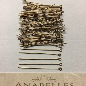 Puede incluir: Un conjunto de alfileres de ojo dorados, ideales para la creación de joyas, dispuestos sobre un fondo claro. Los alfileres son delgados, con un extremo en bucle para sujetar cuentas u otros componentes. La imagen también muestra una parte de una etiqueta vintage de abalorios y hallazgos.