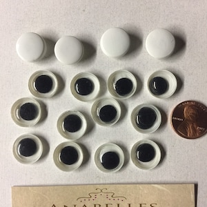 May include: A collection of 12 black and white googly eyes, with a penny for scale. The eyes are clear plastic with black pupils. There are also four white plastic buttons in the image.