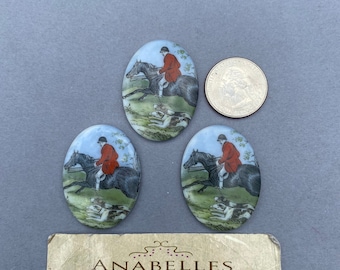 May include: Three oval-shaped ceramic buttons with a vintage illustration of a person riding a horse with hounds. The buttons are white with a blue background and have a glossy finish. The buttons are approximately 1 inch in diameter.