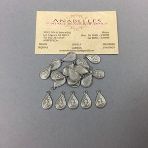 May include: A pile of silver teardrop-shaped metal charms with a floral design. The charms have a hole at the top for attaching to jewelry or other crafts.