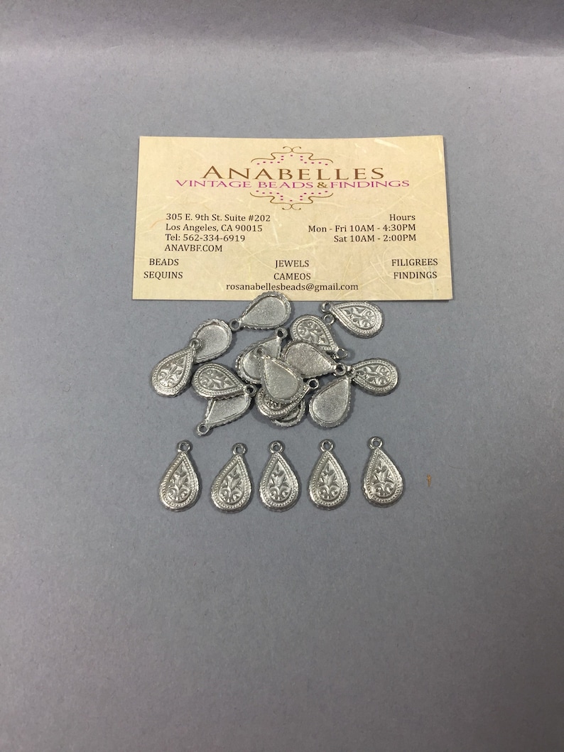 May include: A pile of silver teardrop-shaped metal charms with a floral design. The charms have a hole at the top for attaching to jewelry or other crafts.