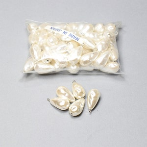 May include: A bag of white pearlescent teardrop-shaped beads with a silver loop at the top. The bag has a label that reads "MADE IN JAPAN". There are also five beads outside of the bag.