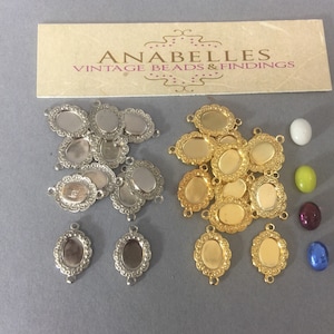 May include: A collection of 10 silver and 10 gold oval-shaped metal bezel settings with a scalloped edge. The bezels are designed to hold a cabochon stone. There are also four colorful cabochon stones in the image: a white, a green, a purple, and a blue.