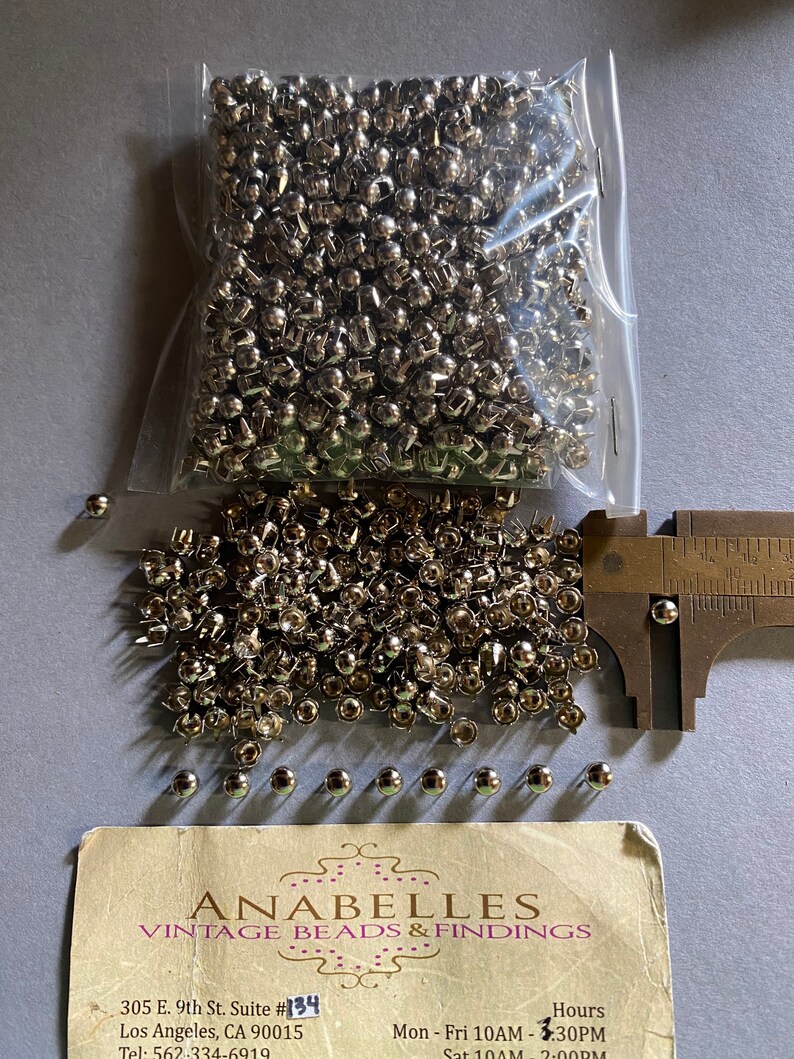 May include: A bag of silver metal rivets, with a few scattered on a white surface. A ruler is in the background. The image includes a business card for Anabelles Vintage Beads & Findings, with the address 305 E. 9th St. Suite #134 Los Angeles, CA 90015, Tel: 562-334-6919, and opening hours.