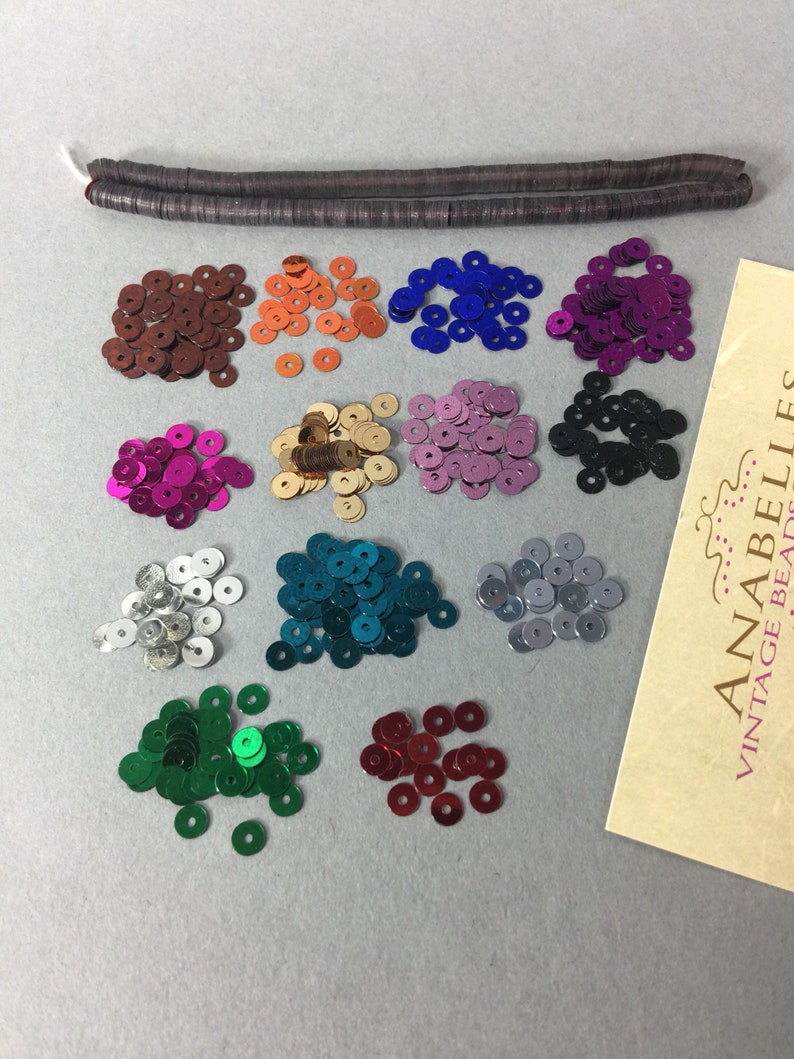 May include: Assortment of 12 colors of round sequins, each color in a separate pile. The sequins are metallic and have a shiny finish. The sequins are arranged in rows on a light gray surface. A brown cord is in the top of the image. A card with the text "ANABELLES... VINTAGE BEADS" is in the bottom right corner of the image.