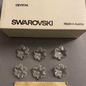 May include: Six clear crystal snowflake charms, each measuring 25 mm, packaged in a white box with the text "ART. 6707", "CRYSTAL", "SWAROVSKI", "2/12 GROSS", "SPECIAL", "25 MM", and "Made in Austria".