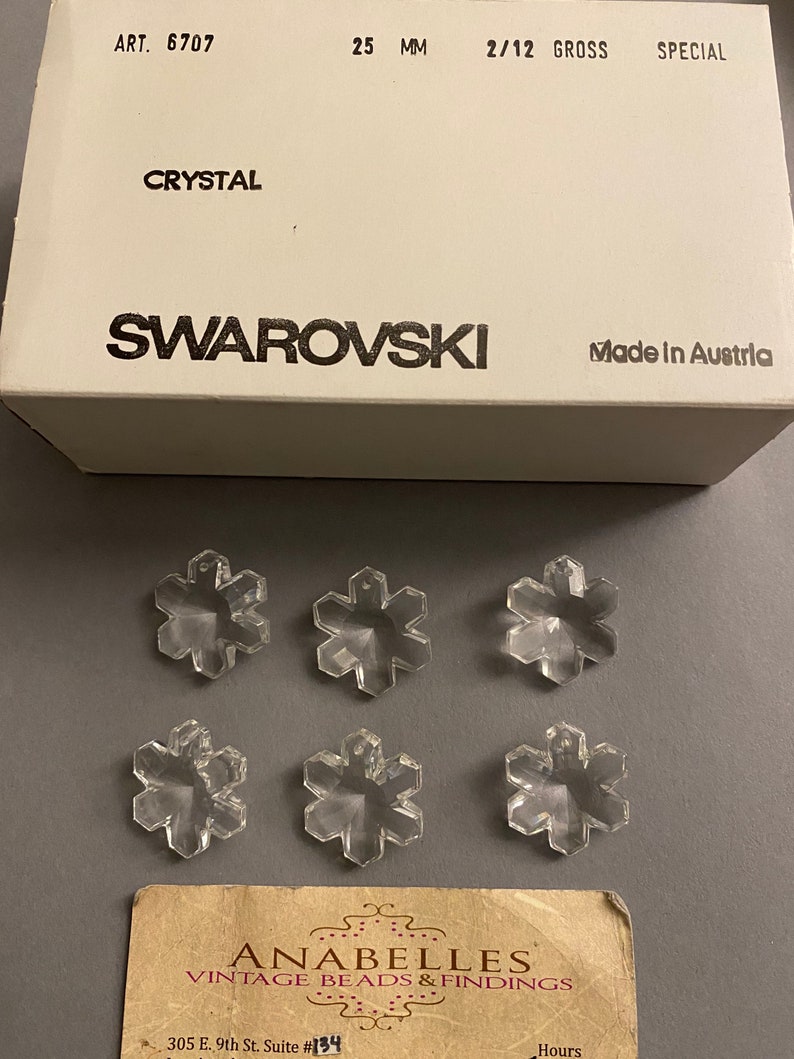 May include: Six clear crystal snowflake charms, each measuring 25 mm, packaged in a white box with the text "ART. 6707", "CRYSTAL", "SWAROVSKI", "2/12 GROSS", "SPECIAL", "25 MM", and "Made in Austria".