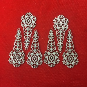 May include: Six silver filigree metal embellishments with a circular design in the center and a teardrop shape on either side. The embellishments are on a red background.