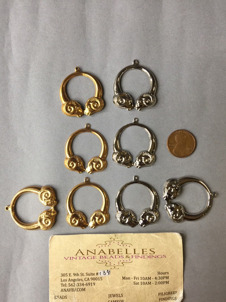 May include: A collection of vintage metal ram head charms in gold and silver tones. The charms are circular with a ram head design on each side. A penny is included for size reference. A business card is also present.