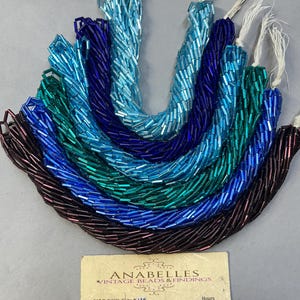 May include: A collection of colorful glass seed beads in shades of blue, green, and purple. The beads are strung together, forming multiple strands. A business card with the name "ANABELLES VINTAGE BEADS & FINDINGS" is in the foreground.