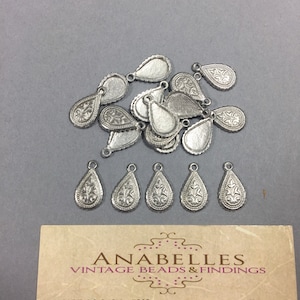 May include: A pile of 16 silver teardrop-shaped metal charms with a decorative floral design. The charms have a hole at the top for attaching to jewelry or other crafts.