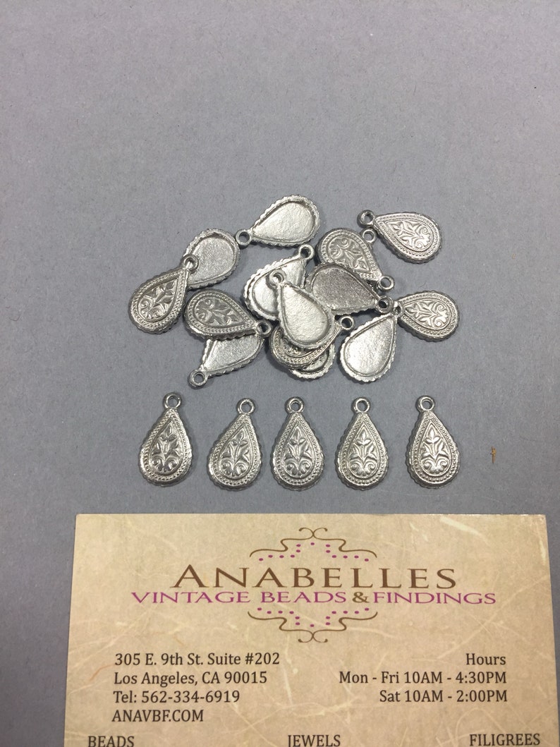 May include: A pile of 16 silver teardrop-shaped metal charms with a decorative floral design. The charms have a hole at the top for attaching to jewelry or other crafts.